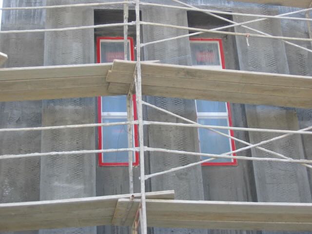 a building under construction with the beginnings of stucco applied, with red tape securing visquine over the windows